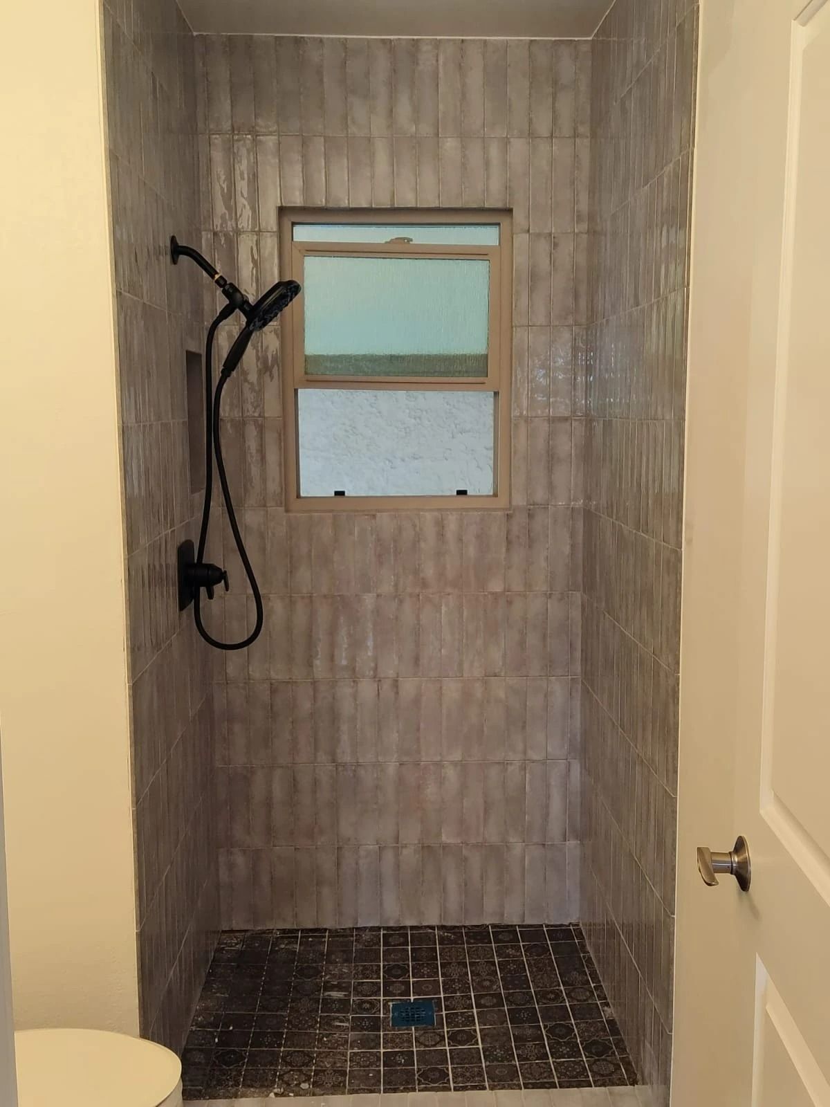 Small tiled shower stall with black shower fixture, window, and dark floor tiles.