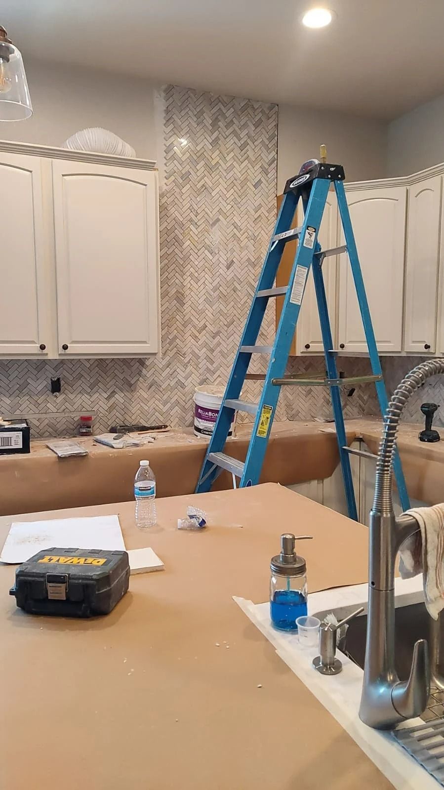 Kitchen under renovation with blue ladder, cabinets, countertop tools, and backsplash tiles in progress