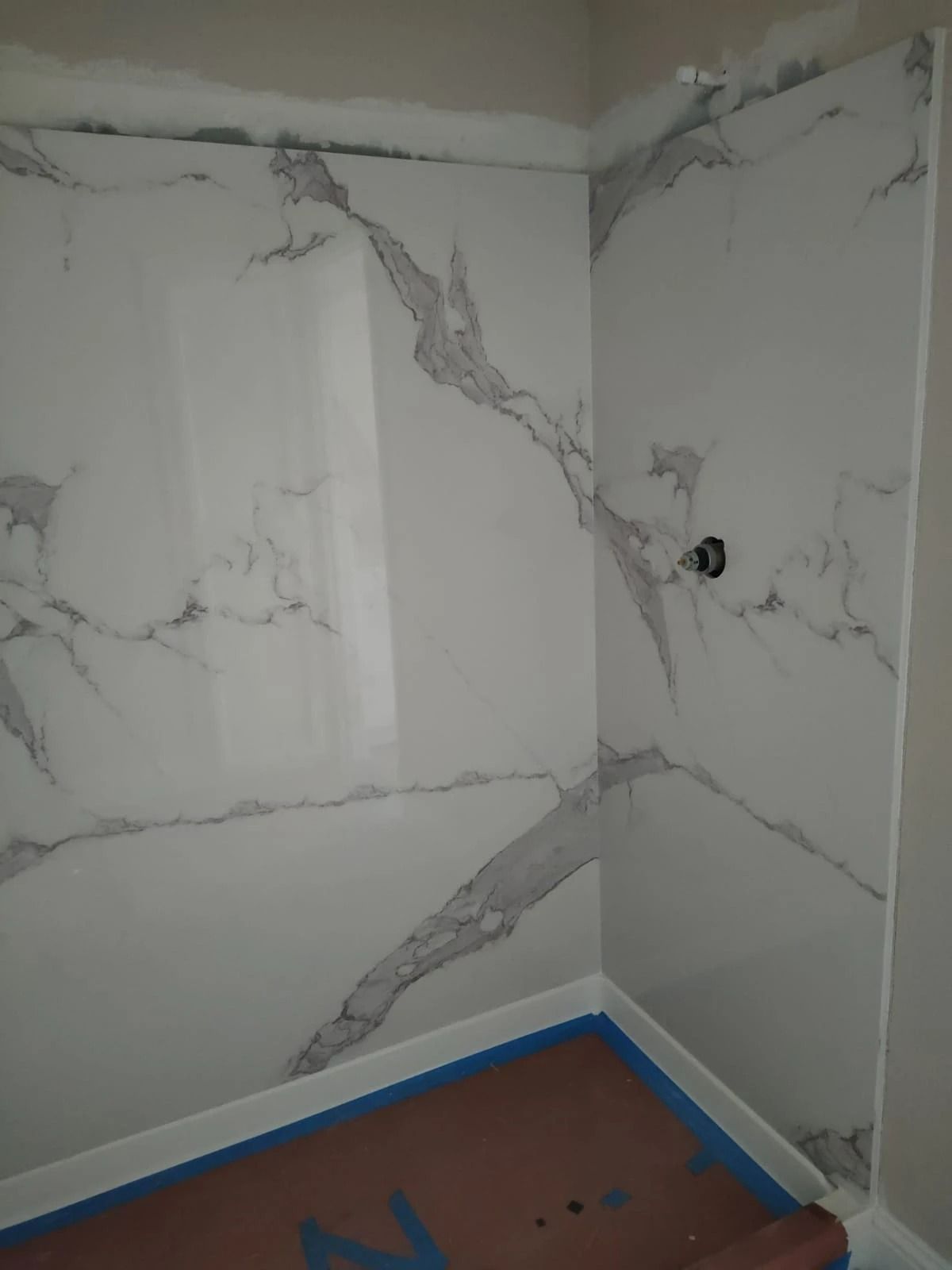 Empty corner of a room with gray marble-pattern wall panels and a brown floor protected by blue tape.