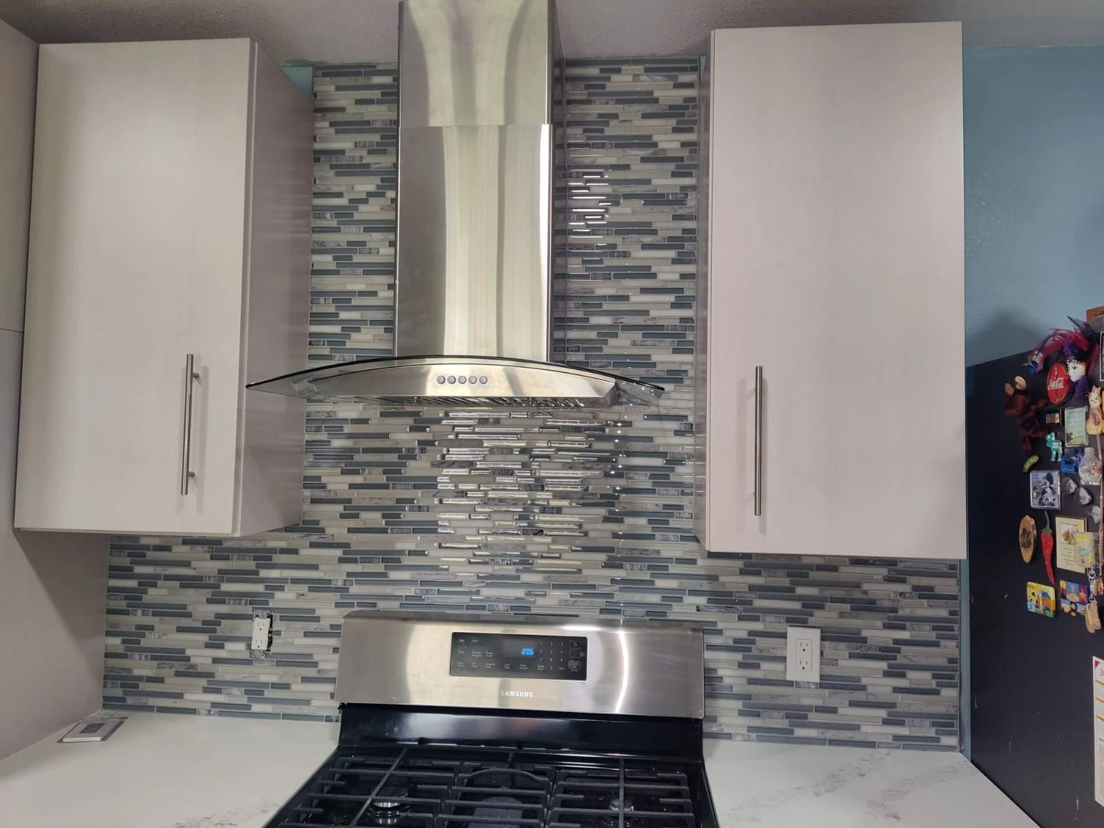 Modern kitchen stove with stainless steel hood, gray mosaic backsplash, and white cabinets
