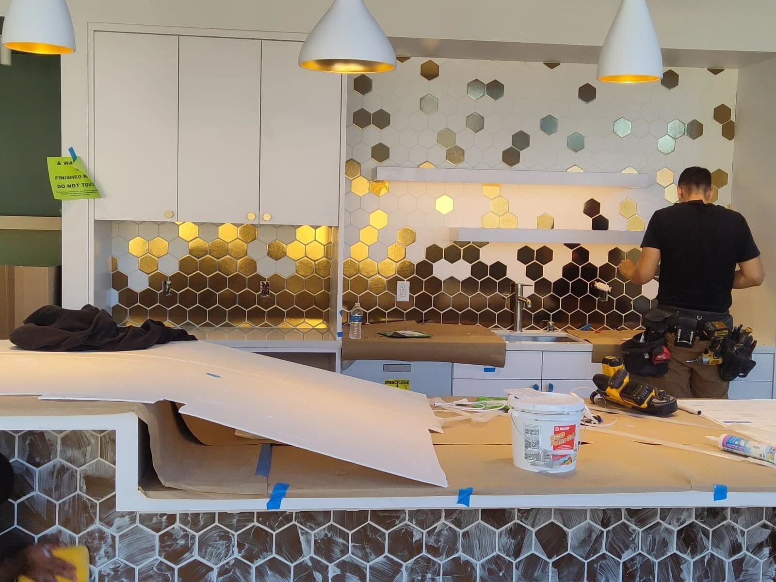Person working at a cluttered counter in a modern kitchen with yellow pendant lights and hexagon tile wall.