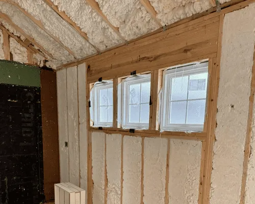 Interior view of a room under construction with white spray foam insulation on the walls and ceiling and three windows.