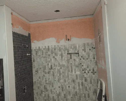 A shower under renovation with partially installed gray rectangular tiles and a dark tiled side wall, against peach walls.