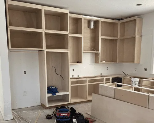 Unfinished light-colored wooden kitchen cabinets installed along walls with a center island in a room under construction.