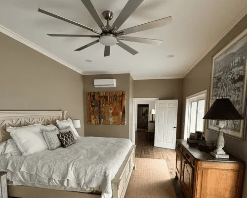 A modern bedroom with a large ceiling fan, a king bed with white linens, a wooden dresser, and artwork on neutral walls.