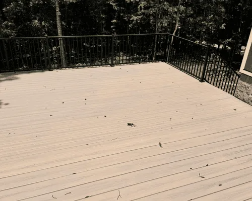Light-colored wood composite deck with black metal railing, surrounded by trees, scattered with dry leaves.