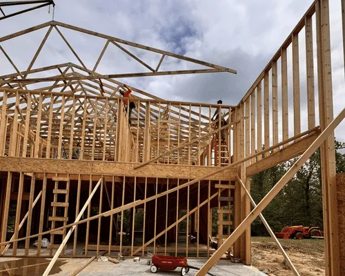 Construction workers framing the walls and installing roof trusses on a wood-framed house under a cloudy sky.
