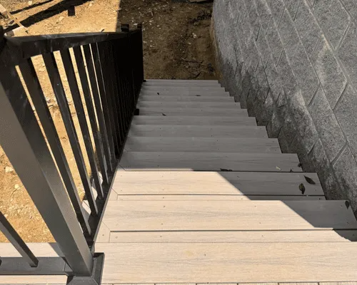A top-down view of a set of light gray composite stairs with a black metal railing next to a gray concrete wall.
