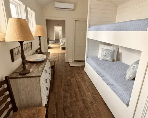 A room featuring a white dresser with two lamps, next to a built-in bunk bed with blue patterned bedding and wooden floors.