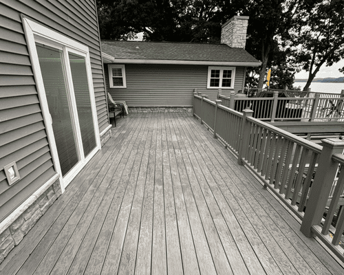 A wooden deck overlooking a lake, featuring a railing, a sliding glass door, and a gray house exterior.