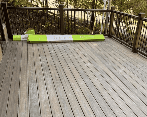 A wooden deck with a black railing, featuring a long, green cardboard package lying on the floor surface.
