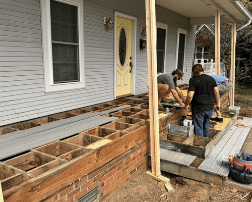 Two construction workers installing deck boards on a porch frame attached to a house with gray siding and a yellow door.