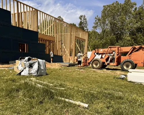 Construction site with a partially framed wooden building, a person standing nearby, and an orange telehandler.