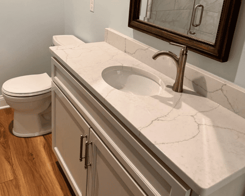 A modern bathroom vanity with a white marbled countertop, bronze faucet, white cabinet, and a nearby toilet.