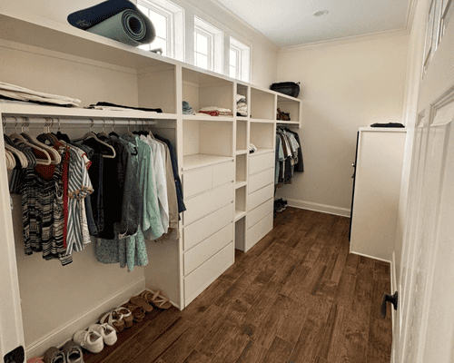 A walk-in closet featuring white built-in shelving, drawers, hanging clothes, and wood-look flooring under three windows.