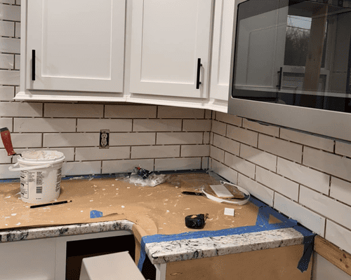 A kitchen backsplash being installed with white subway tiles, a bucket of mortar, and painter's tape on the countertops.