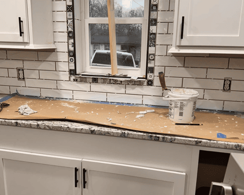 White subway tile backsplash partially installed on a kitchen wall around a window with granite countertops and cabinets.