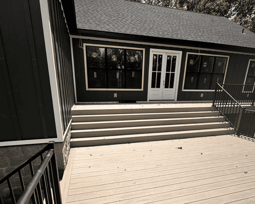 A dark-sided house with white double doors, wide steps leading up to a deck, and black railings on a sunny day.