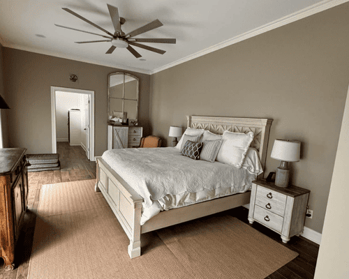 A bedroom with a light-colored wood bed frame, beige walls, white bedding, ceiling fan, and area rug.