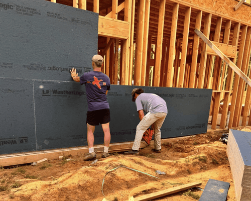 Two workers install dark grey structural sheathing panels onto the wooden frame of a building under construction.
