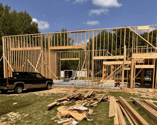 A black and tan pickup truck parked in front of a wooden building frame under construction on a sunny, grassy lot.