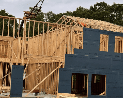 A multi-story house under construction with exposed wooden framing and blue sheathing panels against a backdrop of trees.