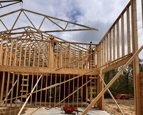 Construction workers frame the wooden walls and roof trusses of a house under a cloudy sky.