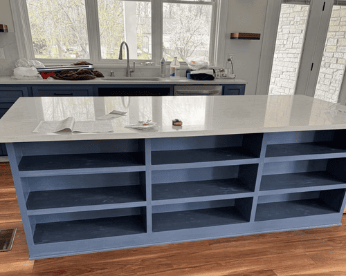 A large blue kitchen island with open shelving and a white countertop stands in a brightly lit kitchen.