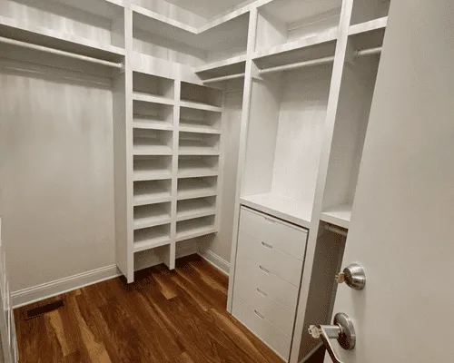 Empty white walk-in closet with built-in shelves, hanging rods, and a four-drawer unit, set on hardwood flooring.