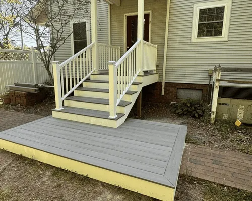 A low, gray wooden deck with matching stairs and white railings leading up to the front entrance of a yellow house.