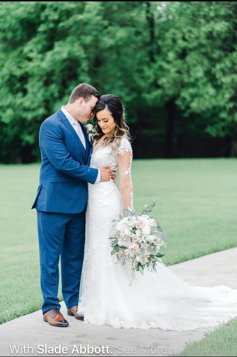 Newlyweds embrace outdoors: Groom in blue suit, bride in lace gown holds bouquet.