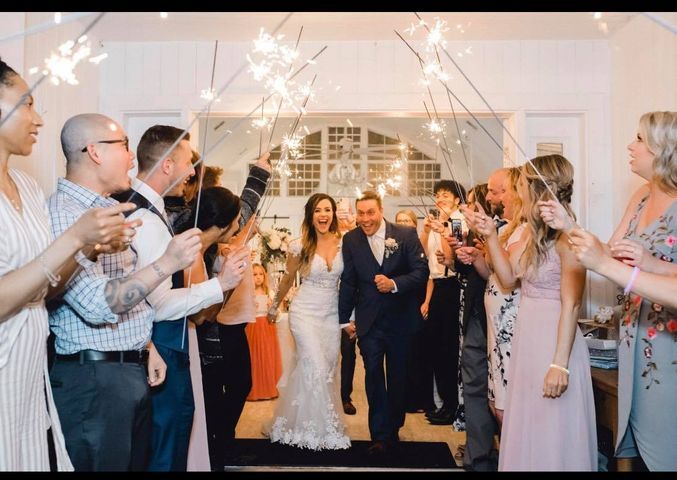 Bride and groom walk under sparklers held by wedding guests, celebrating inside a venue.