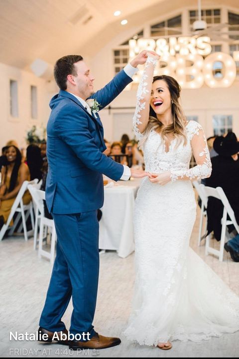 Bride and groom dancing at a wedding reception. She wears a white dress, he wears a blue suit; both smile.