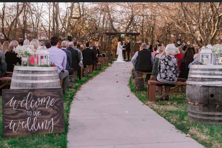 Outdoor wedding ceremony; couple at altar, guests seated on benches, barrels with flowers and sign.