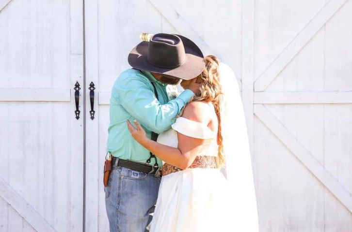 Couple kissing in front of white barn doors. The man wears a cowboy hat and the woman a wedding dress.