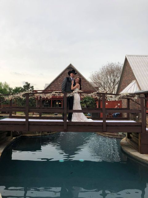 Couple in wedding attire on a bridge over a pool, buildings in the background. Overcast sky.