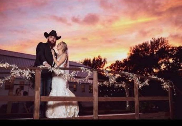 Couple on a wooden bridge, gazing at sunset. Groom in cowboy hat, bride in white dress.