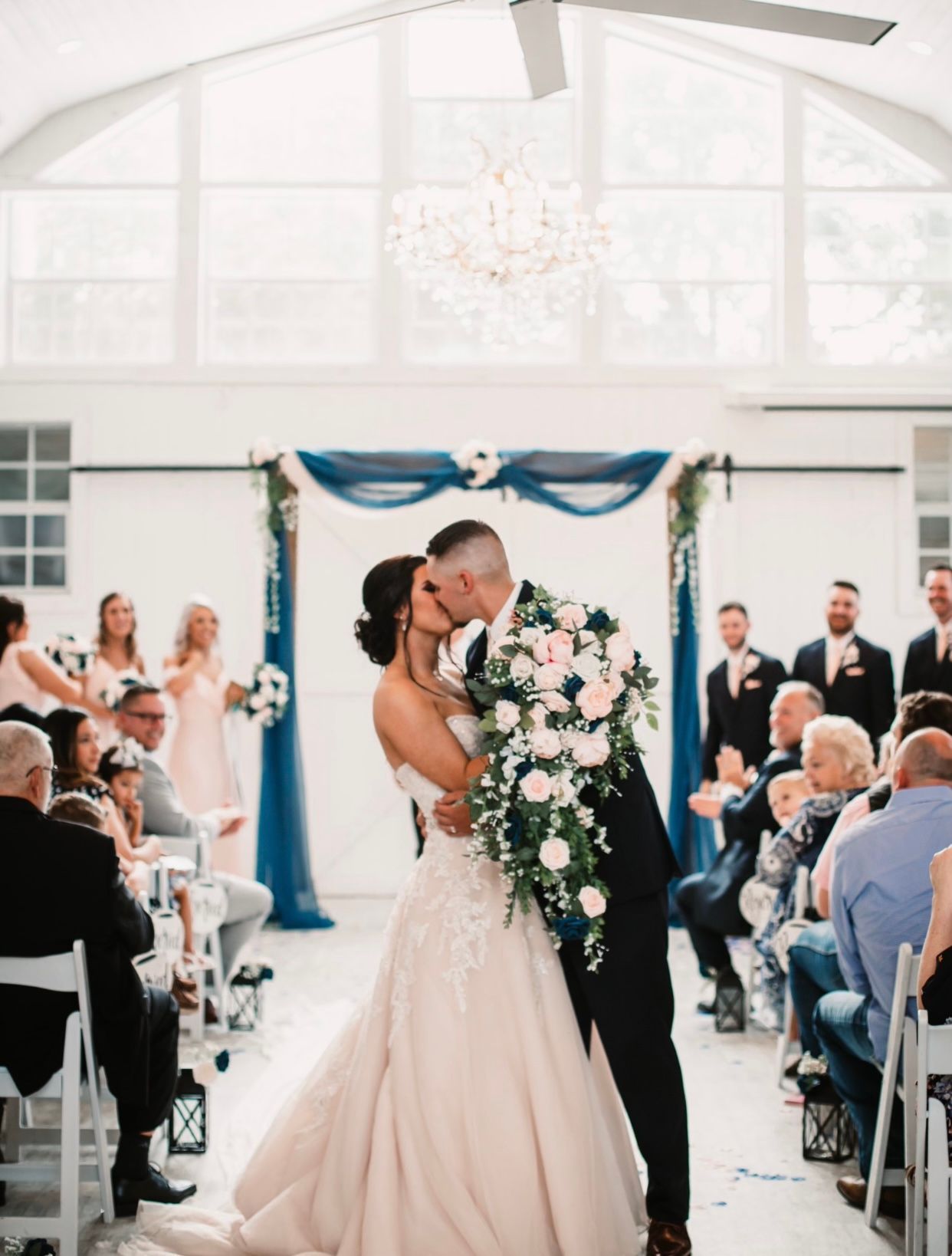 Bride and groom kissing at altar; wedding ceremony with guests and bridesmaids, blue and white decor.