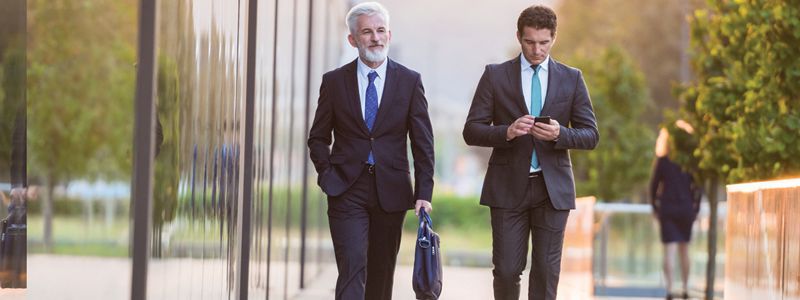 Two businessmen in suits walk outdoors, one carrying a briefcase and the other checking his phone.