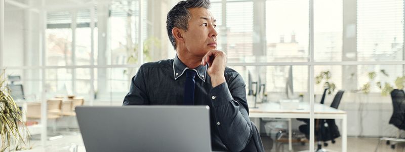 Man in a dark shirt looking pensively out a window in a bright office; laptop in foreground.