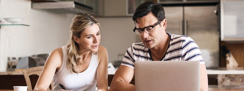 A couple looks at a laptop in a kitchen. The man speaks, and the woman listens intently.