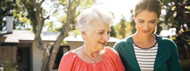 An older woman and a younger woman walk outside together. The older woman smiles while the younger woman looks down, wearing a teal cardigan.
