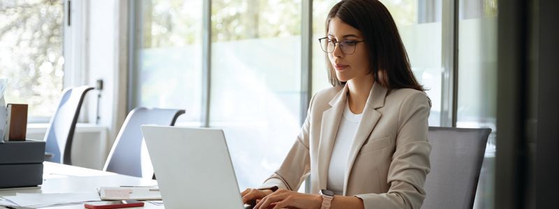 Woman working on a laptop at a bright office desk, wearing glasses and a beige blazer