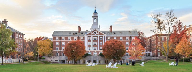 Large brick building with a clock tower surrounded by colorful trees on a green lawn.