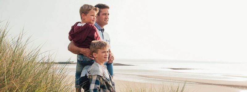 Family standing on a beach, looking out at the water near grassy dunes.