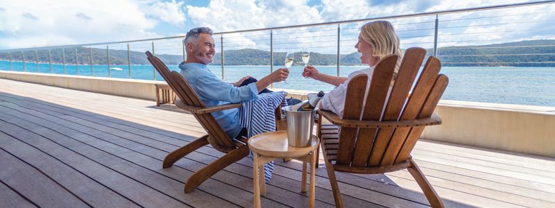 Two people relaxing in wooden chairs on a seaside deck, holding drinks with the ocean in view