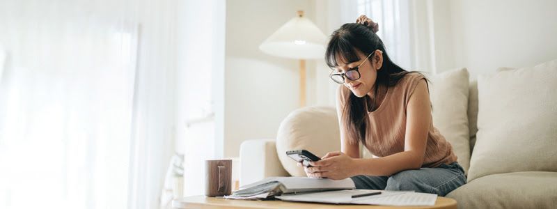 Woman with glasses looking at her phone while sitting on a couch next to paperwork and a speaker in a living room.