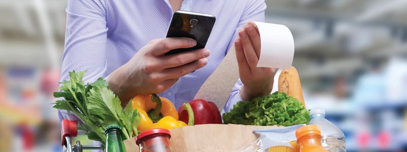 Person holding a phone beside a grocery basket of fresh vegetables and a receipt in a store aisle