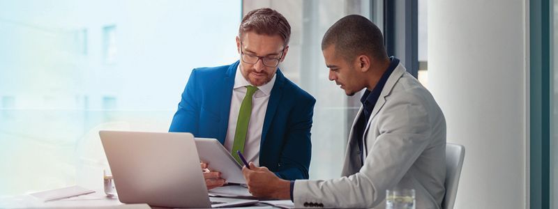 two men looking at a notebook and computer screen