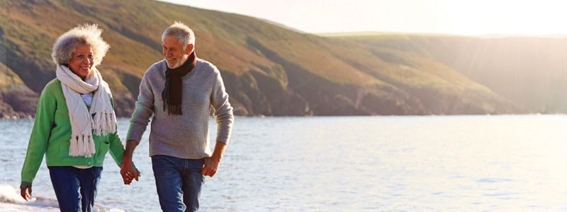 an elderly couple is walking on the beach holding hands.